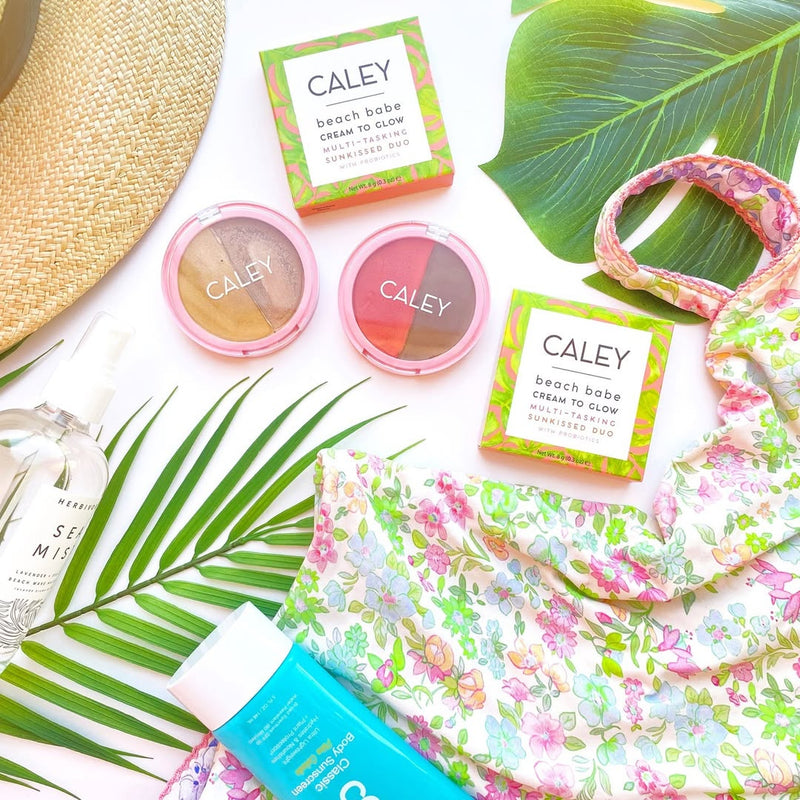 Flat lay of Caley Cosmetics makeup products styled with tropical beach accessories and palm leaves on a bright summer-inspired background.