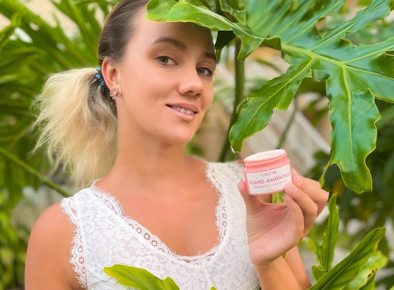 Caley Cosmetics Island Ambrosia moisturizer held outdoors among lush tropical greenery for a fresh, dewy skin moment.