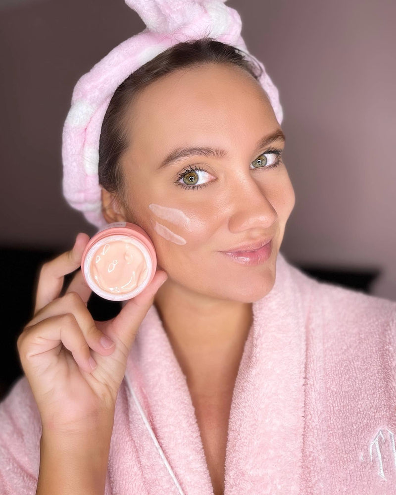 Woman holding Caley Island Ambrosia moisturizer with cream swatches on her cheek, highlighting a probiotic skincare routine for healthy-looking skin.