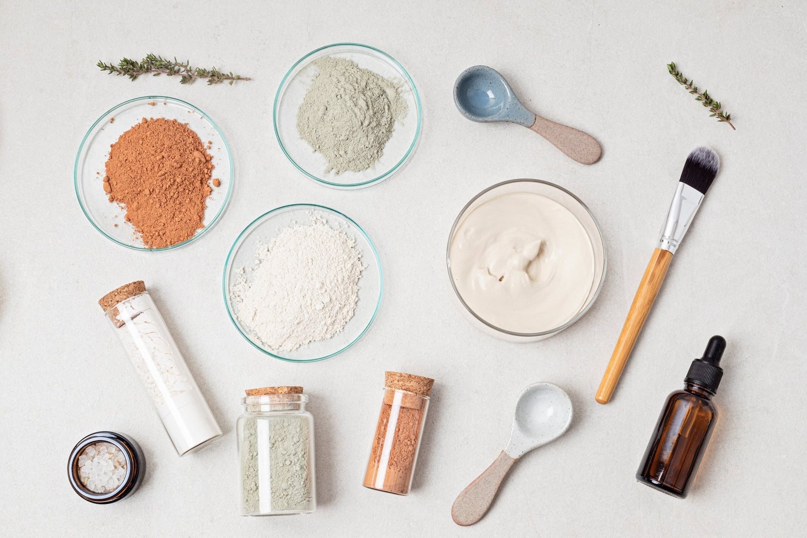 Assorted cosmetic ingredients including powders, cream jars, dropper bottles, and makeup brushes arranged on a clean surface.