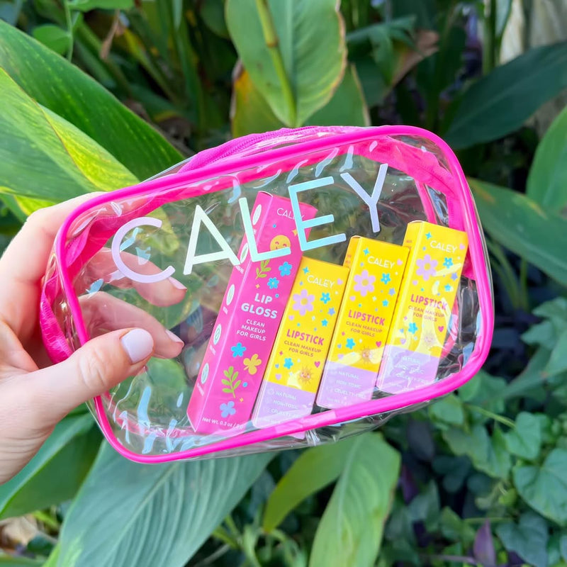 Hand holding Caley Cosmetics Clean and Cute Lip Kit with lipstick and lip gloss against green leaves.