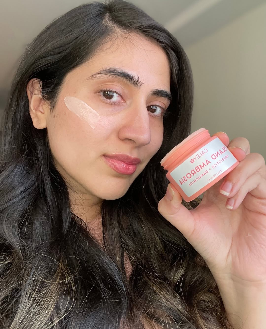 Woman holding a skincare jar near her face with a small swipe of cream on her cheek, highlighting probiotic moisturizer use.