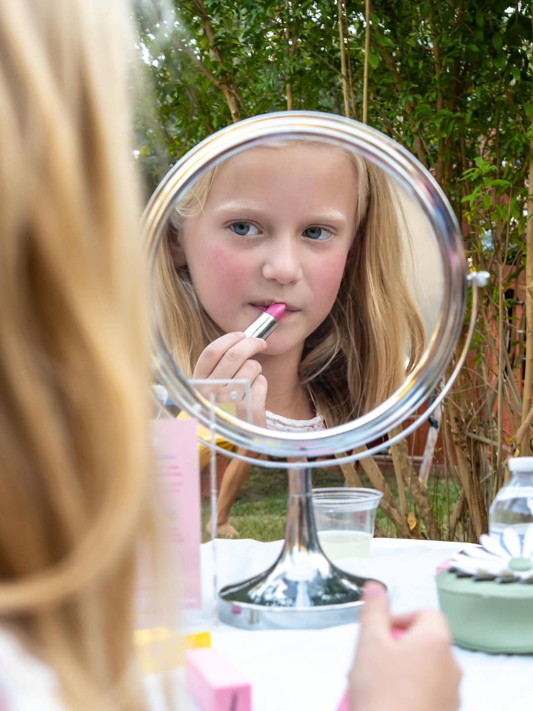Child applying Caley Cosmetics Happy Tint lipstick while looking into a round tabletop mirror outdoors.