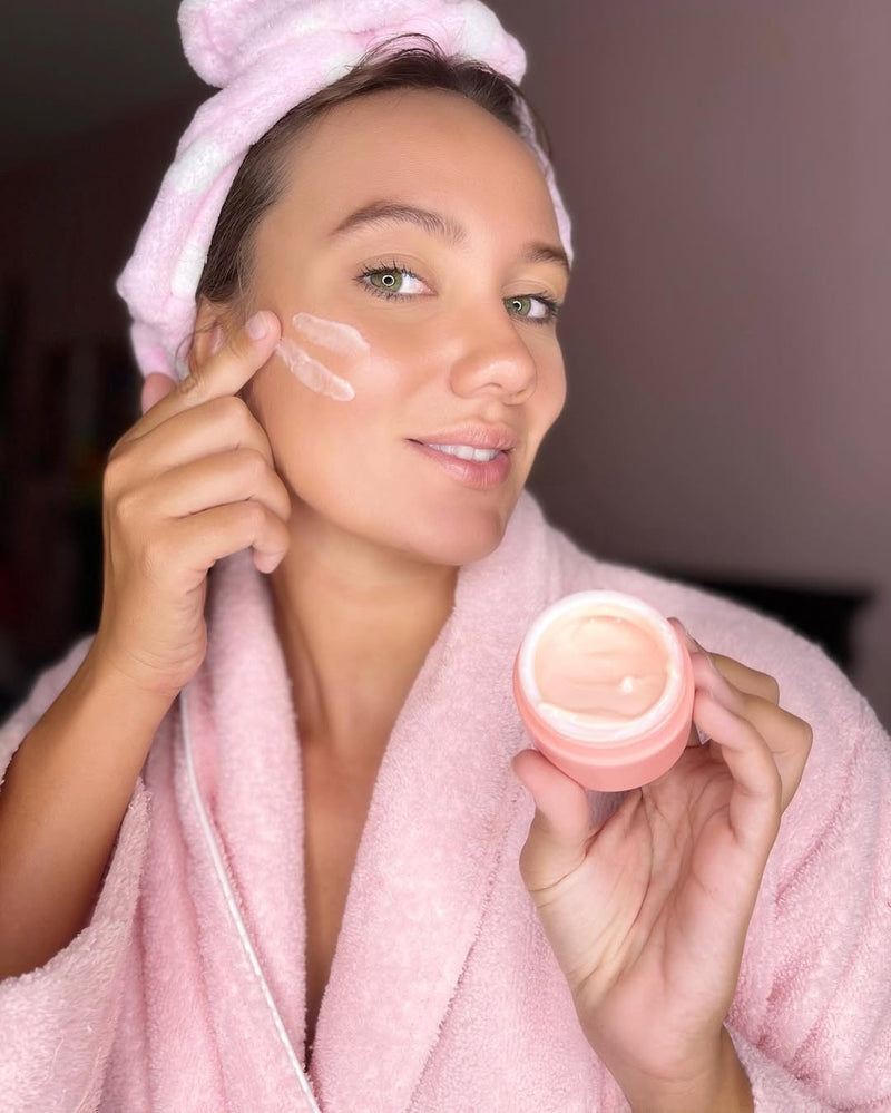 Woman in a pink robe applying Caley Cosmetics Island Ambrosia Bakuchiol Moisturizer during her skincare routine.