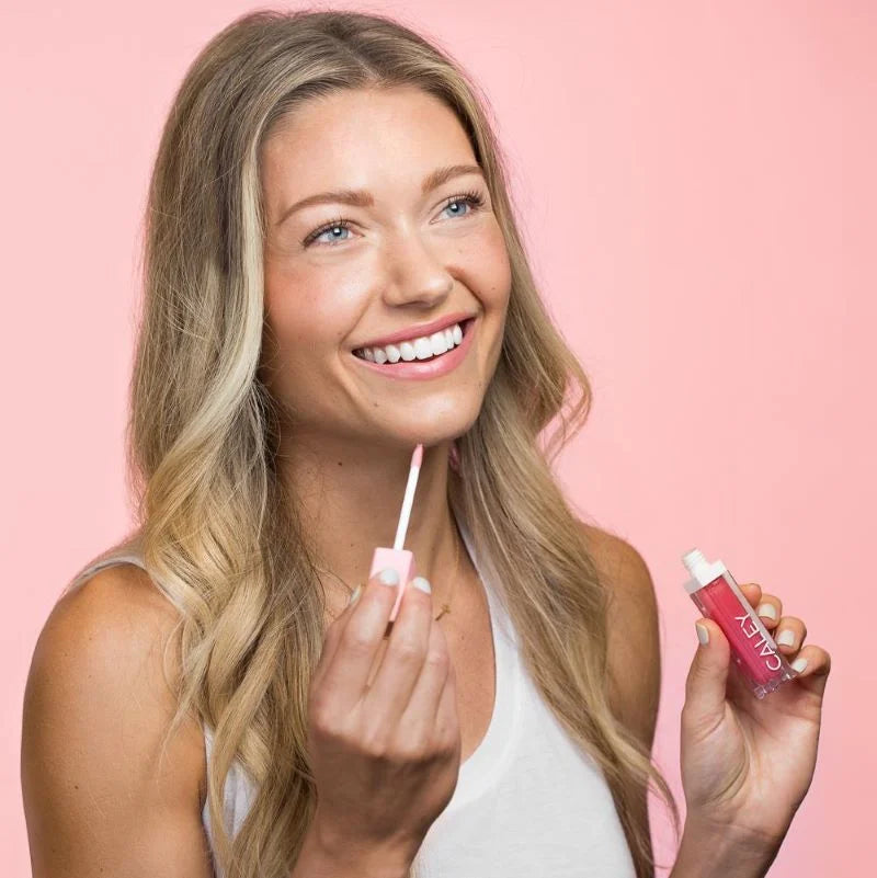 Woman holding Caley lip gloss and applicator against a soft pink background.