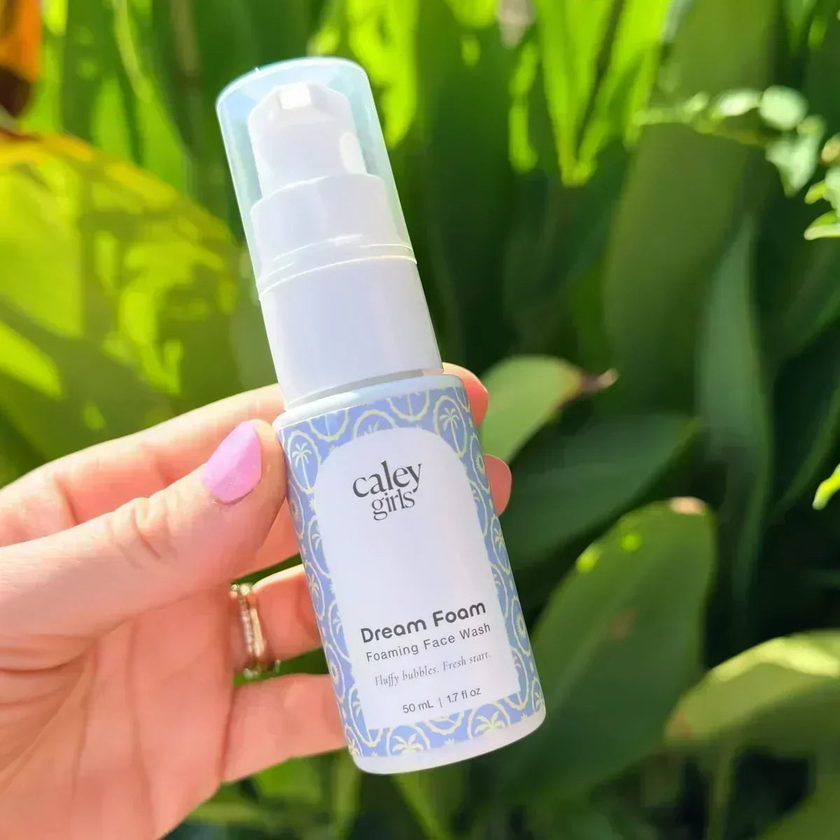 Caley Girls Dream Foam Foaming Face Wash held in sunlight against lush green tropical leaves.