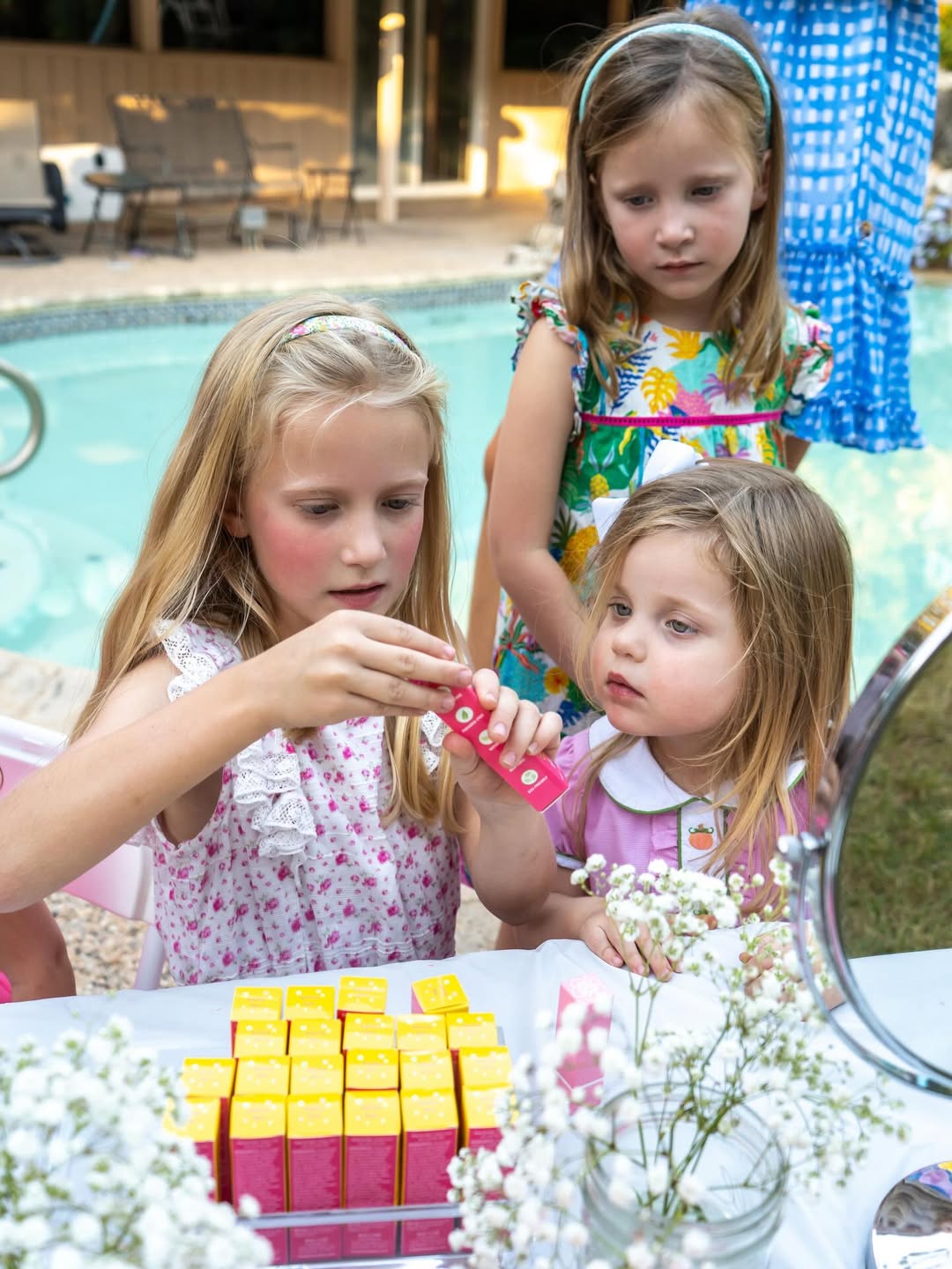 Children gathered around a table, examining Caley Cosmetics lip balm tubes beside a mirror outdoors.