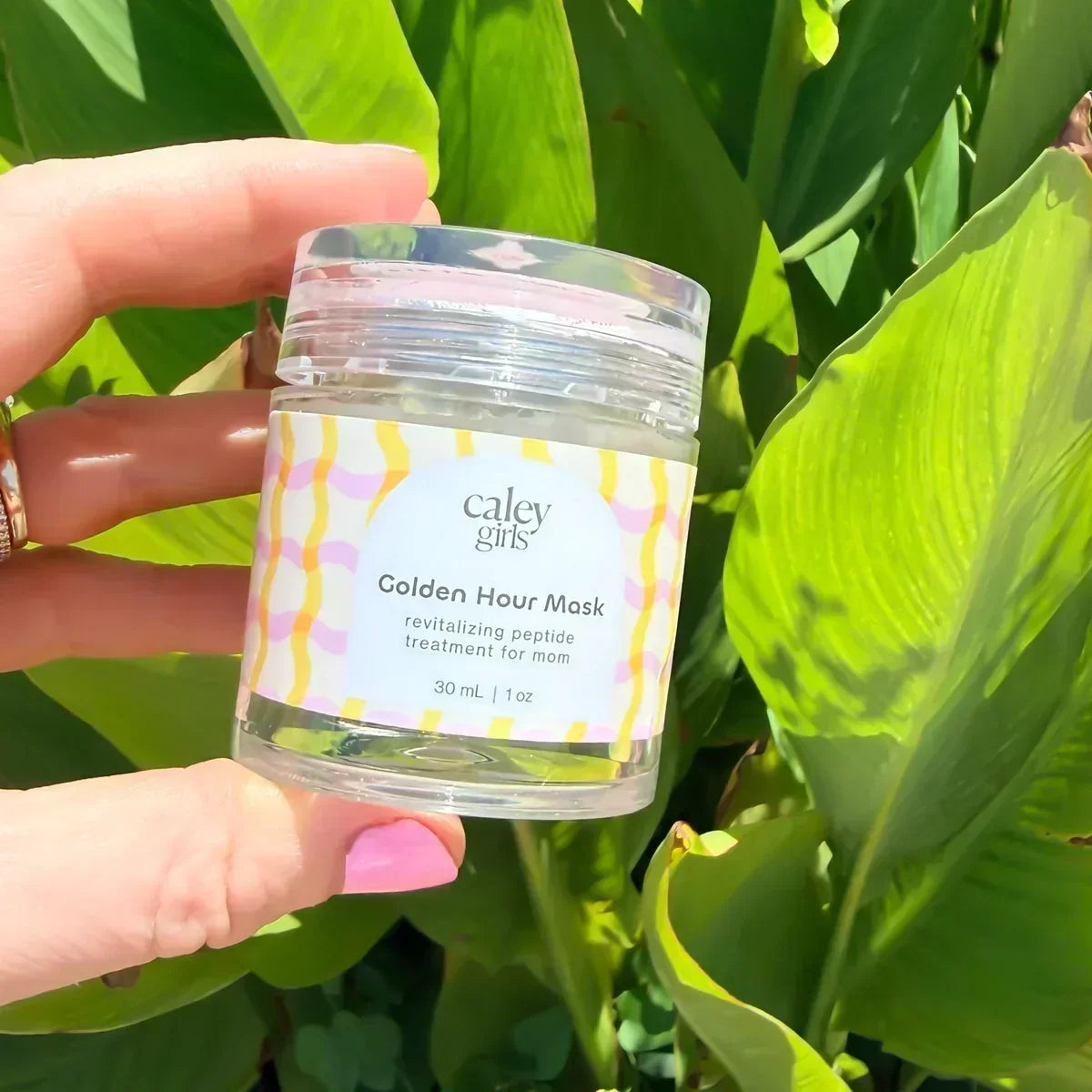 Caley Girls Golden Hour Mask held in sunlight against lush green tropical leaves.
