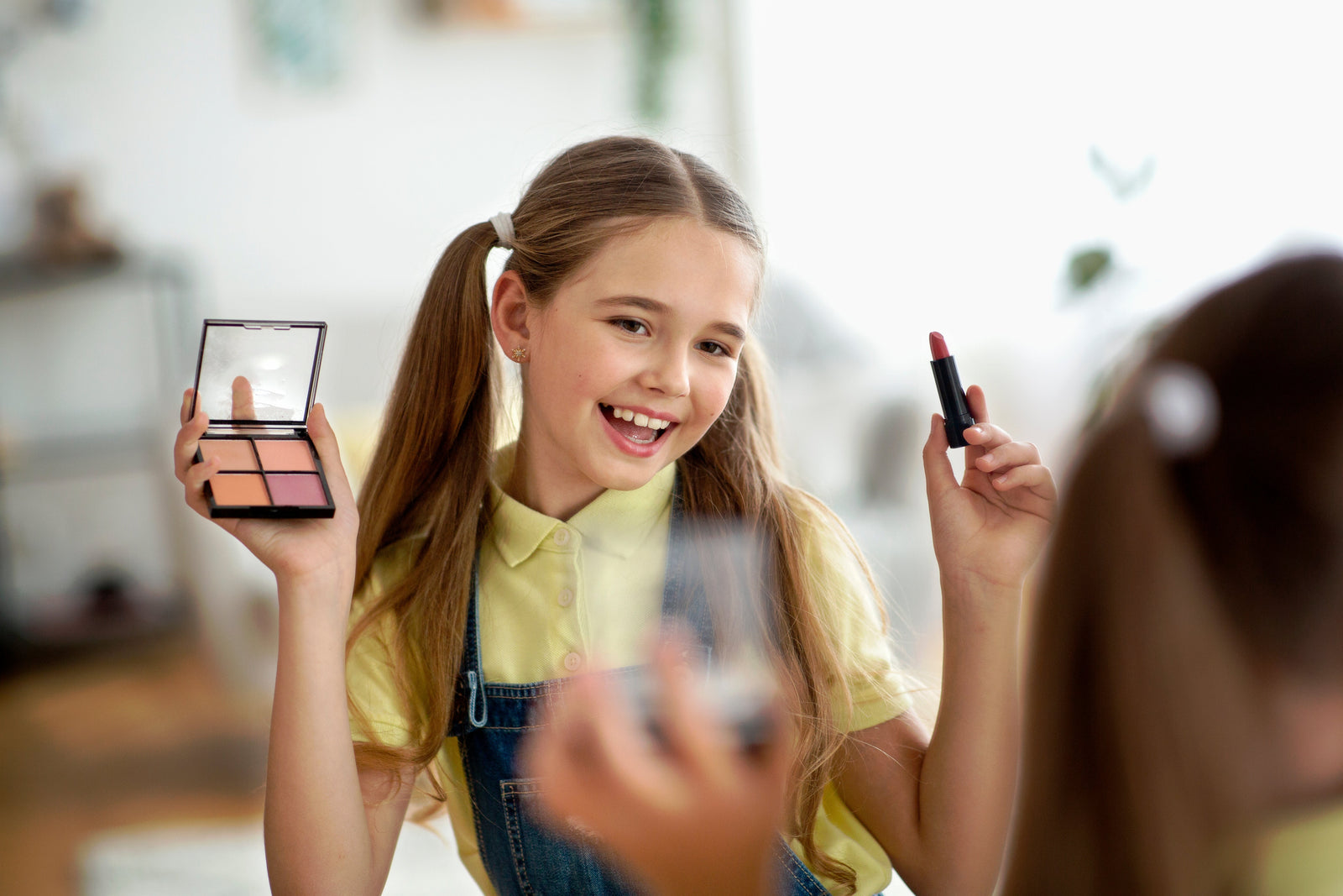 Smiling tween girl with pigtails holding a Caley Cosmetics makeup palette and lipstick while looking in a mirror.